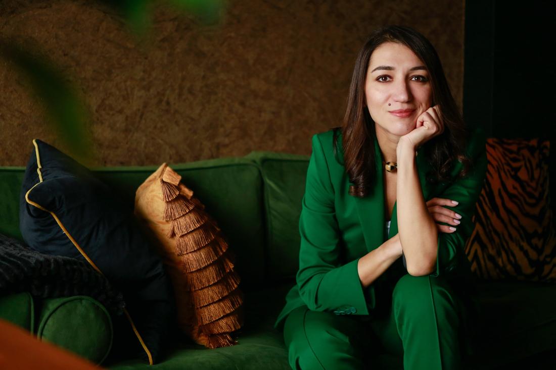 Irina smiling on a green velvet sofa in a green suit